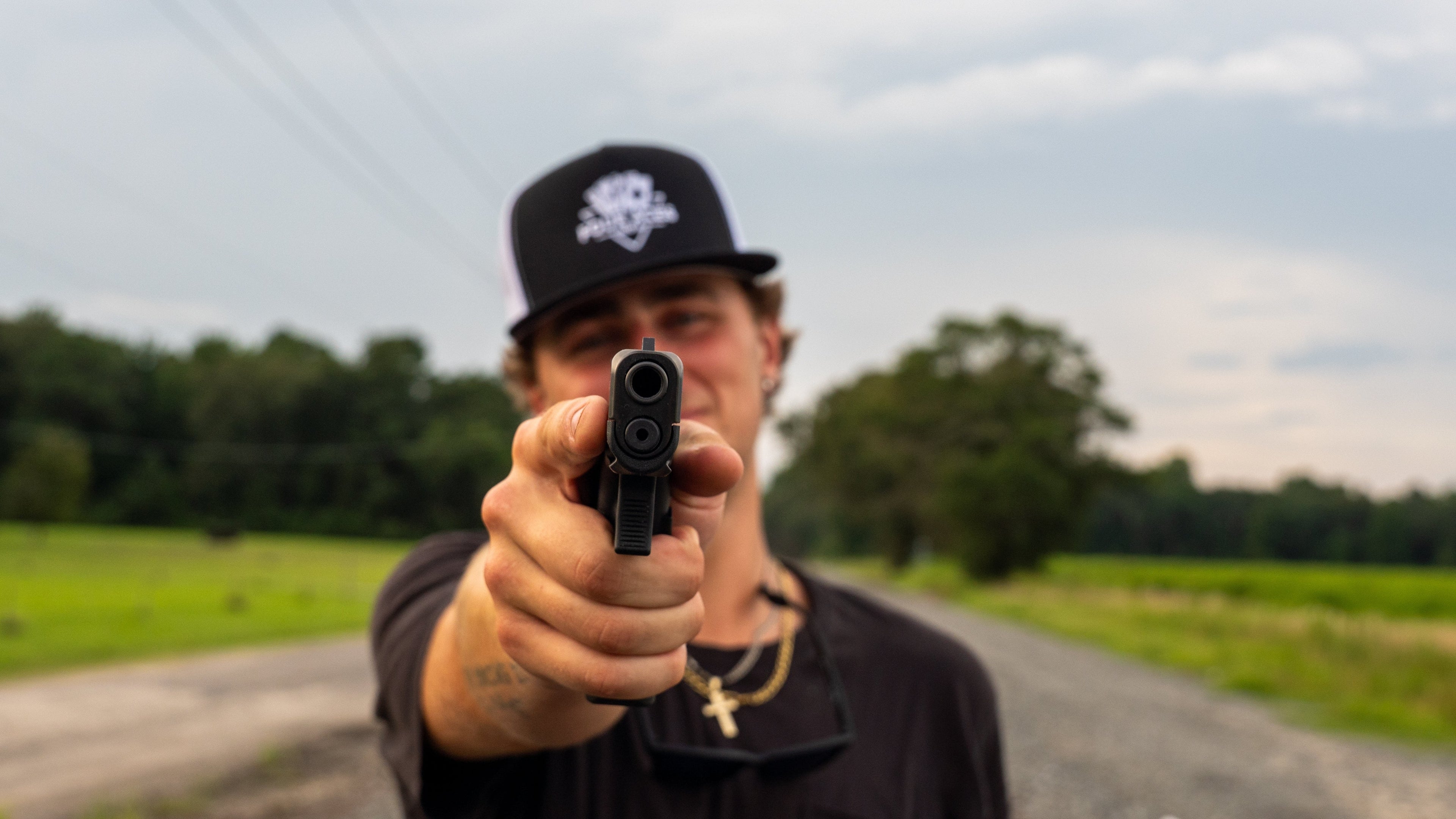 Person holding a handgun on a rural road with trees and sky in the background